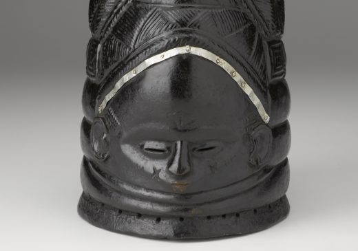 Una máscara negra con un rostro humano y un peinado alto en forma de cúpula tallado en su superficie.