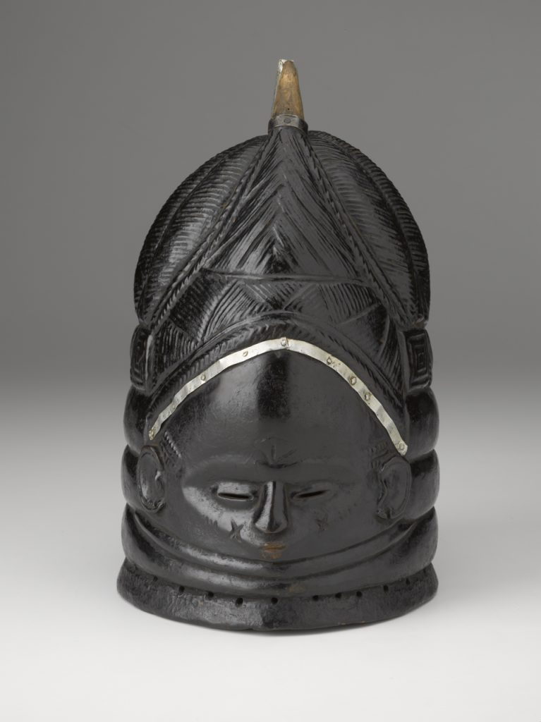 Una máscara negra con un rostro humano y un peinado alto en forma de cúpula tallado en su superficie.
