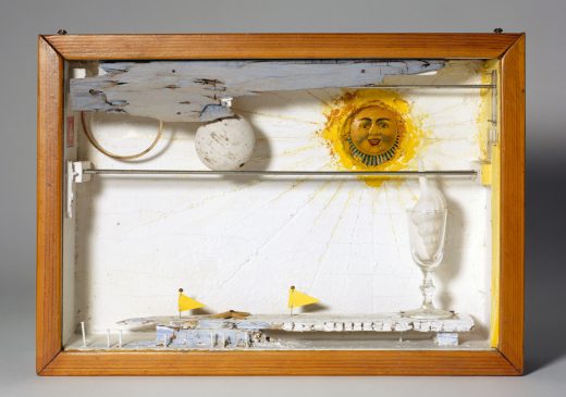 A clear shadow box with a wooden frame around it. Inside the box is a sun, a golf ball, a boat, a piece of driftwood, and a shell inside a glass goblet.