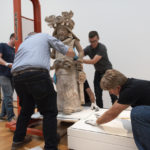 Four people position the sculpture of the female diety onto a pedestal. 