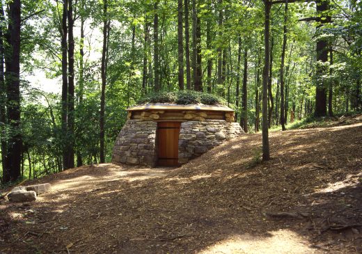 A photograph of a stone hut with green plants growing on top of its wooden roof. The hut is located on a hill and surrounded by tall trees.