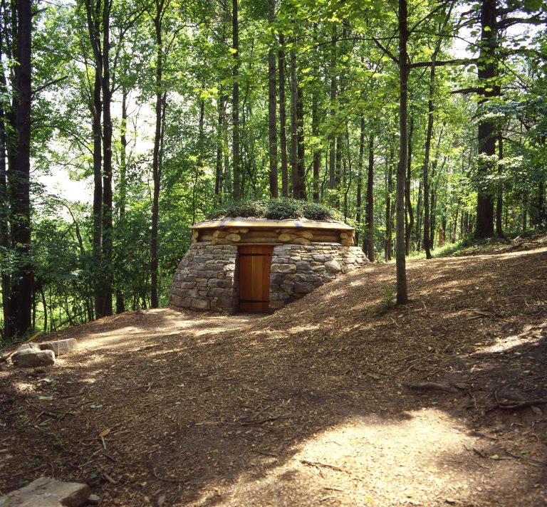 A photograph of a stone hut with green plants growing on top of its wooden roof. The hut is located on a hill and surrounded by tall trees.