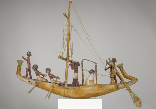 Maqueta de madera de un barco con una tripulación de figuras humanas y un pasajero vestido de blanco.
