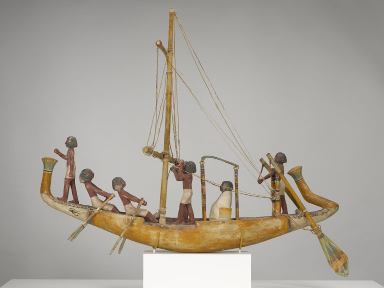 Maqueta de madera de un barco con una tripulación de figuras humanas y un pasajero vestido de blanco.