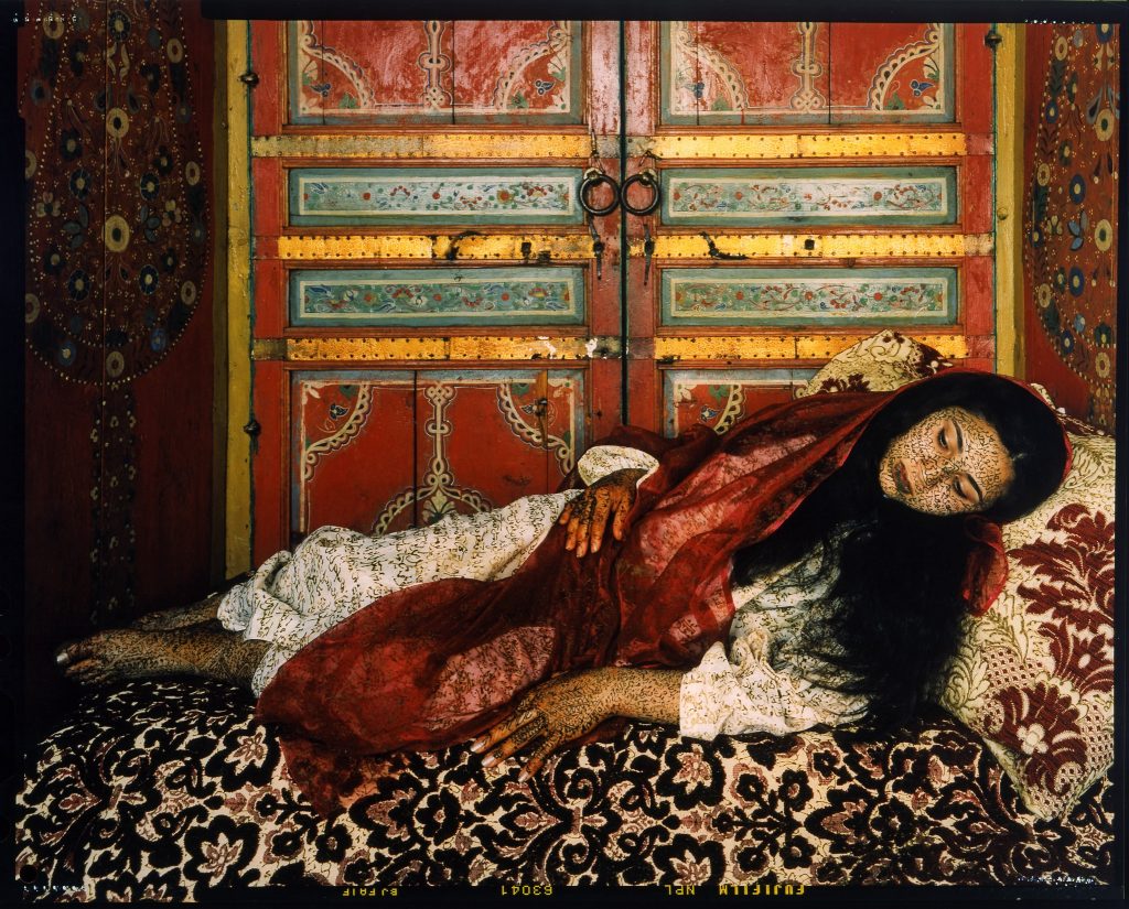 A color photograph of a dark-haired woman lying on a bed covered with floral-patterned fabric. She is wearing a red veil over a white robe, with writing on the fabric and on her skin.