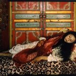 A color photograph of a dark-haired woman lying on a bed covered with floral-patterned fabric. She is wearing a red veil over a white robe, with writing on the fabric and on her skin.