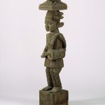 A carved wooden post made up of two human figures stacked one on top of the other and separated by a patterned geometric form. The figure at the top has an elaborate hairstyle and holds a staff in each hand. The figure at the bottom wears a hat and holds a drum in one hand.