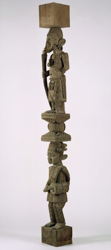 Poste de madera tallada compuesto por dos figuras humanas apiladas una sobre otra y separadas por una forma geométrica estampada. La figura de arriba lleva un elaborado peinado y sostiene un bastón en cada mano. La figura de abajo lleva un sombrero y sostiene un tambor en una mano.