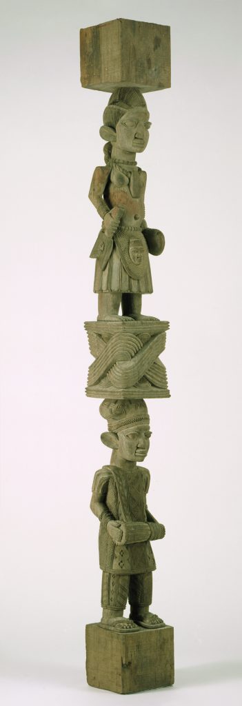 A carved wooden post made up of two human figures stacked one on top of the other and separated by a patterned geometric form. The figure at the top has an elaborate hairstyle, wears necklaces and a skirt with a face on it, and holds ritual objects in each hand. The figure at the bottom holds a drum and wears a hat and a diamond-patterned outfit.