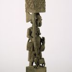 A carved wooden post made up of two human figures stacked one on top of the other and separated by a patterned geometric form. The figure at the top has an elaborate hairstyle and protruding breasts, and she holds a ritual object with a face on it. The figure at the bottom holds a dead bird in one hand and an ax in the other. A dog stands at his feet.