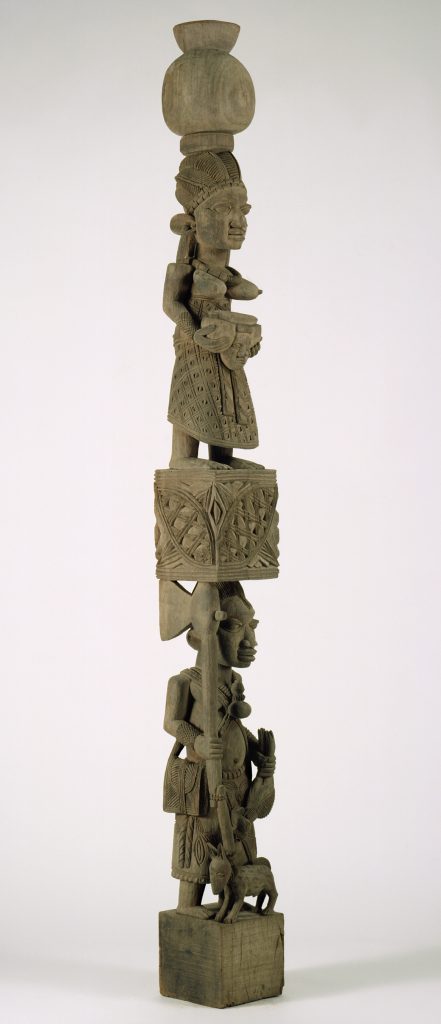 A carved wooden post made up of two human figures stacked one on top of the other and separated by a patterned geometric form. The figure at the top has an elaborate hairstyle and protruding breasts, and she holds a ritual object with a face on it. The figure at the bottom holds a dead bird in one hand and an ax in the other. A dog stands at his feet.