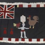 Una bandera de tela pintada con imágenes rojas, blancas y azul oscuro de una bandera británica en una esquina y un hombre en el centro sosteniendo dos grandes pájaros y balanceando un reloj sobre su cabeza.