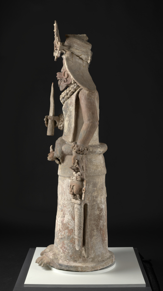 Escultura de una deidad femenina que sostiene una antorcha en la mano derecha y una bolsa en la izquierda.