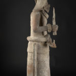Side view of a ceramic sculpture of a female holding a torch in her right hand and a bag in her left hand.