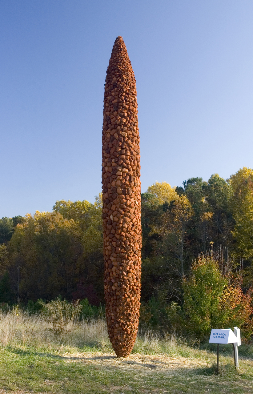 MarthaJacksonJarvis.jpg A tall, textured, red-orange sculpture on a grassy hill, with tall trees and a blue sky in the background. The sculpture has a cylinder shape that tapers to a point at the top and the bottom.