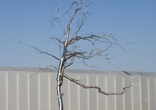 A silver tree-like sculpture on a grassy hill. The sculpture has a thick trunk and thin, barren branches. There is a white building and a blue sky in the background.