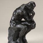 A bronze sculpture of nude male figure seated on a rock, propping his face on his hand and appearing to be deep in thought.