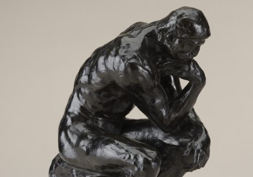 A bronze sculpture of nude male figure seated on a rock, propping his face on his hand and appearing to be deep in thought.
