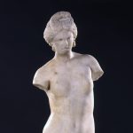 Estatua de mármol de una figura de diosa desnuda, a la que le faltan los dos brazos, con una figura de delfín a sus pies.