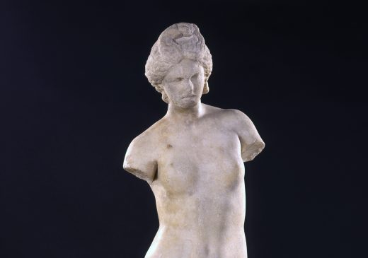 Estatua de mármol de una figura de diosa desnuda, a la que le faltan los dos brazos, con una figura de delfín a sus pies.