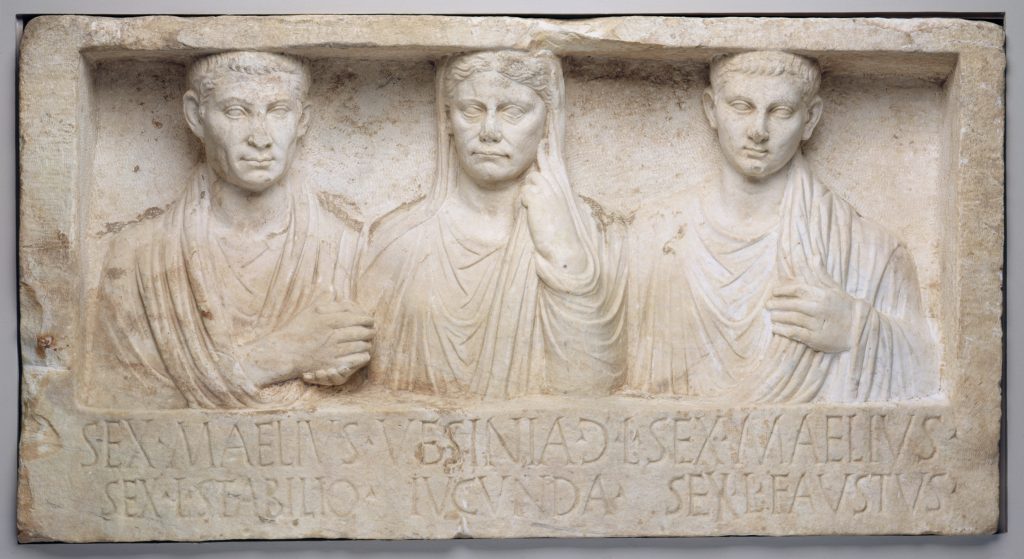 A rectangular, sculpted white marble monument depicting three human figures: a husband and wife and their son. Their names are inscribed in Latin at the bottom.