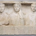 Monumento rectangular de mármol blanco esculpido que representa a tres figuras humanas: un matrimonio y su hijo. Sus nombres están inscritos en latín en la parte inferior.