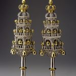 Two matching silver Torah finials, each topped with a gold crown and small gold bells that hang from intricate decorative elements.