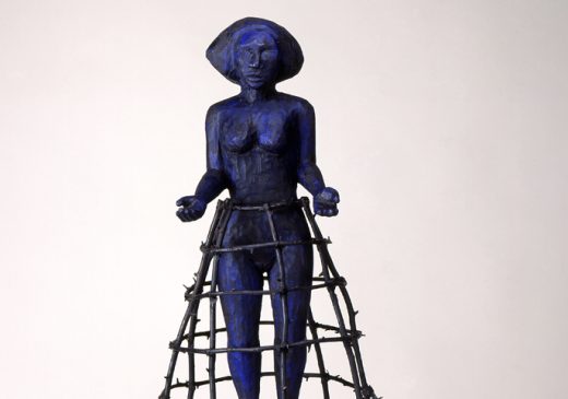 A blue-and-black wooden figure of a woman hovering in the air with her arms outstretched. She is enclosed by a bronze cage that extends from her waist to the floor.