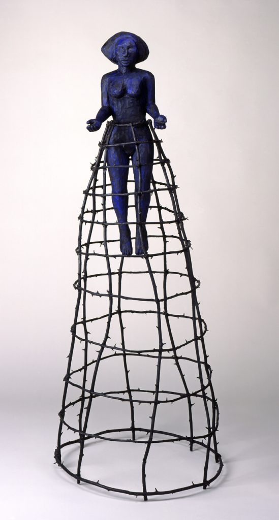 A blue-and-black wooden figure of a woman hovering in the air with her arms outstretched. She is enclosed by a bronze cage that extends from her waist to the floor.