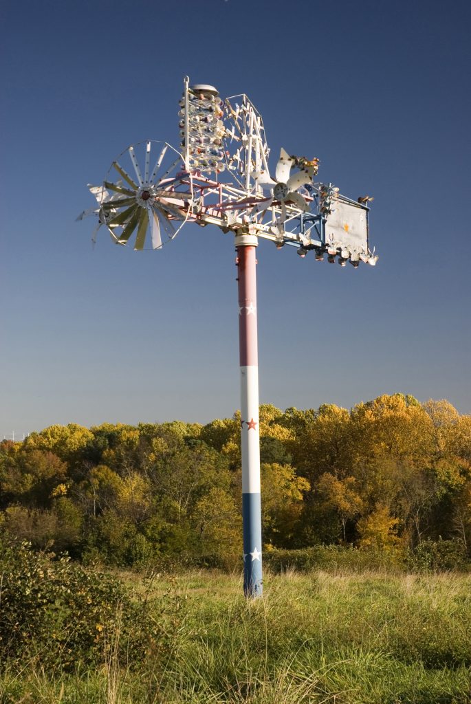A metal sculpture on a grassy hillside, with tall trees and a blue sky in the background. The sculpture has a thick pole that is painted red, white, and blue, with a star design. There are multiple metal pinwheels and other movable parts attached to the top of the pole.