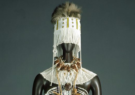 A costume featuring a white wrap skirt trimmed in black, with arm bands, a patterned and beaded torso, necklaces, and strips of animal skin hanging from the hips. The costume includes a headpiece with fur on top and a beaded veil below.