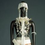 A costume made up of a white wrap skirt trimmed in black stripes, arm bands, a fringed, beaded belt, a fringed headpiece, hoop earrings, a collar tufted with blond hair, and various necklaces dotted with shells.