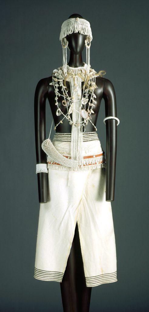 A costume made up of a white wrap skirt trimmed in black stripes, arm bands, a fringed, beaded belt, a fringed headpiece, hoop earrings, a collar tufted with blond hair, and various necklaces dotted with shells.
