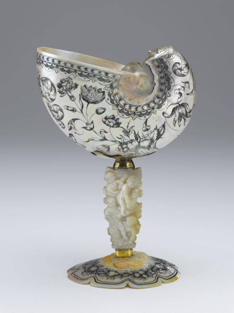 A decorative drinking cup made from an Indonesian nautilus shell. The body of the cup is decorated with a floral pattern. The cup’s stem is a carved oyster shell that is decorated with cupid figures and images of fruit.