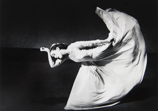 A black-and-white photograph of a woman wearing a long, white dress. She is leaning forward, with the top of her head resting against her wrist and kicking her leg up behind her, creating an arc shape with the bottom half of her dress.