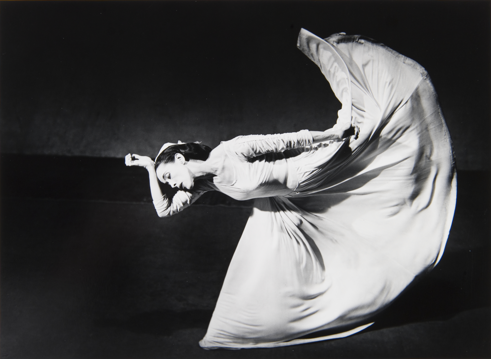 Martha Graham, “Letter to the World” (Kick) Fotografía en blanco y negro de una mujer con un vestido largo y blanco. Está inclinada hacia delante, con la parte superior de la cabeza apoyada en la muñeca y levantando la pierna por detrás, creando una forma de arco con la mitad inferior del vestido.