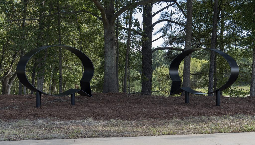 Two large black sculptures of the outlines of speech bubbles in an outdoor setting. There is pine straw on the ground below and surrounding the sculptures. There are tall trees in the background and a sidewalk in the foreground.