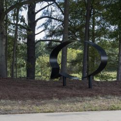 Two large black sculptures of the outlines of speech bubbles in an outdoor setting. There is pine straw on the ground below and surrounding the sculptures. There are tall trees in the background and a sidewalk in the foreground.