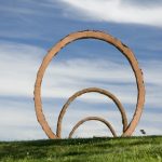 Three large, red-orange, ring-shaped sculptures on a grassy lawn. There is a paved path between two of the rings. In the background there are trees and a blue sky with white, puffy clouds. View through all three rings lined up.
