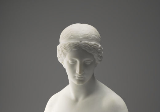 A white marble sculpture of a female figure’s head, neck, shoulders, and chest. Her hair is tied back into a bun, and she is gazing downward. A wreath of laurel leaves surrounds her bare chest.