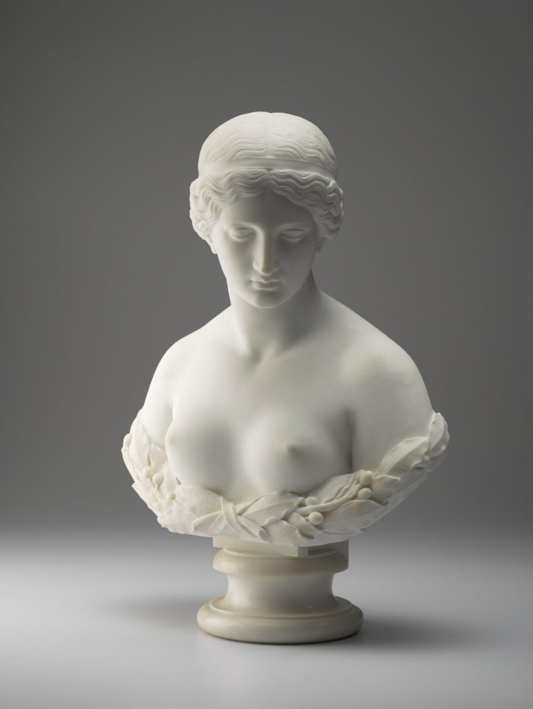 A white marble sculpture of a female figure’s head, neck, shoulders, and chest. Her hair is tied back into a bun, and she is gazing downward. A wreath of laurel leaves surrounds her bare chest.