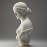 A white marble sculpture of a female figure’s head, neck, shoulders, and chest. Her hair is tied back into a bun, and she is gazing downward. A wreath of laurel leaves surrounds her bare chest. Side View.