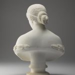 A white marble sculpture of a female figure’s head, neck, shoulders, and chest. Her hair is tied back into a bun, and she is gazing downward. A wreath of laurel leaves surrounds her bare chest. Viewed from the back.