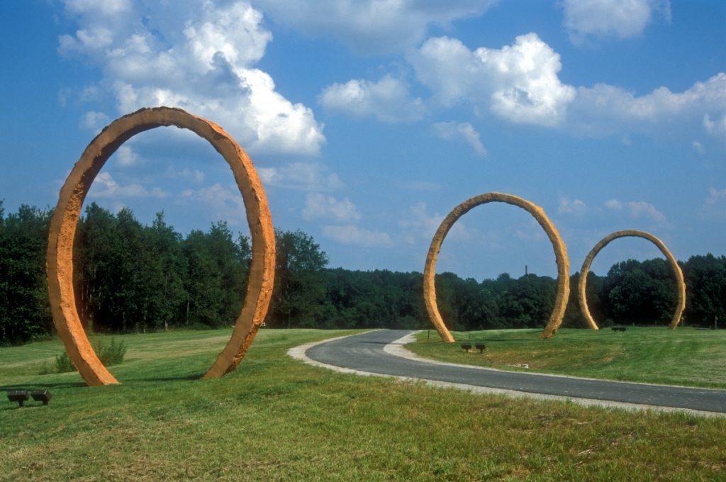 Three large, red-orange, ring-shaped sculptures on a grassy lawn. There is a paved path between two of the rings. In the background there are trees and a blue sky with white, puffy clouds.
