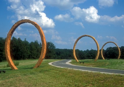 Three large, red-orange, ring-shaped sculptures on a grassy lawn. There is a paved path between two of the rings. In the background there are trees and a blue sky with white, puffy clouds.