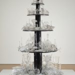 A sculpture made up of five round tiers of black-painted wood displaying hundreds of glass objects that range from completely intact at the top to progressively broken and shattered on each lower level.