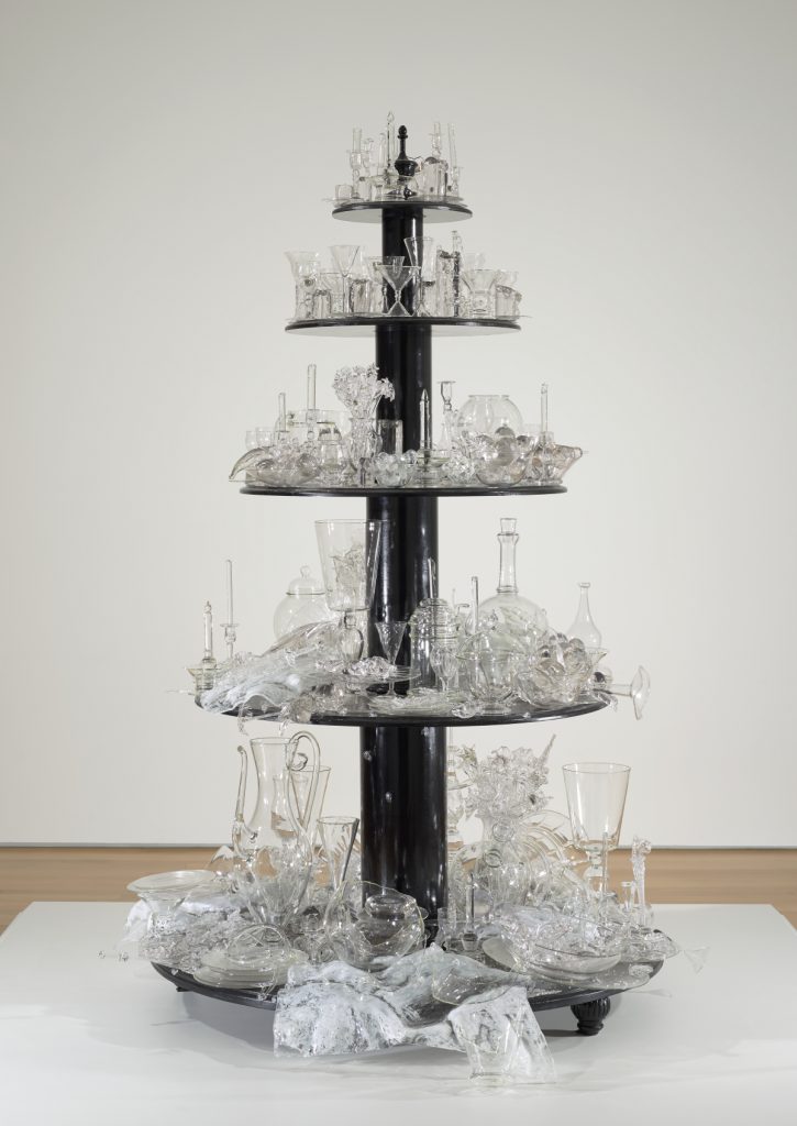 A sculpture made up of five round tiers of black-painted wood displaying hundreds of glass objects that range from completely intact at the top to progressively broken and shattered on each lower level.