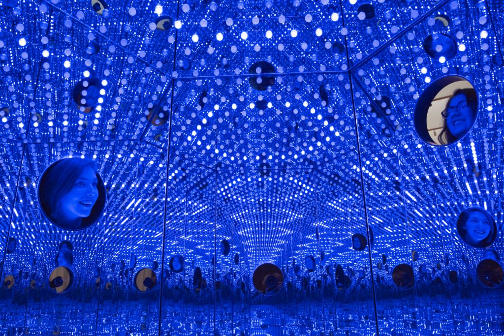 An interior view of a large, hexagon-shaped box filled with hundreds of color-changing light bulbs that reflect off the mirrored walls inside.