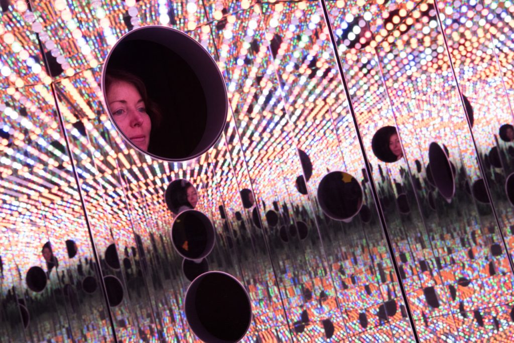 Una vista interior de una gran caja en forma de hexágono llena de cientos de bombillas que cambian de color y se reflejan en las paredes de espejo del interior.