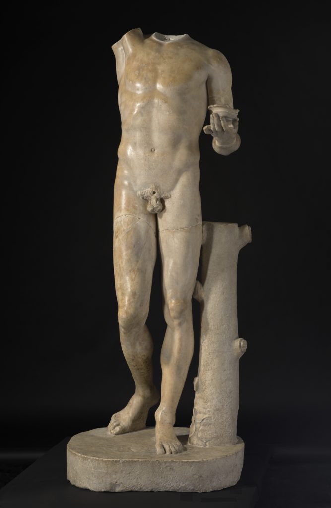 A white marble statue of a nude male figure wearing a crown and holding a bunch of grapes above his head with one hand and a cup in the other.
